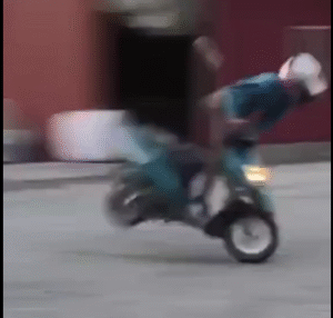 The video begins with a person in a helmet and blue jacket riding a small scooter. They are in an open space featuring a reddish building and several large white bales in the background, all set against an overcast sky.